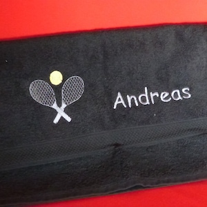Könnte beinhalten: Schwarzes Handtuch mit gestickten Tennisschlägern und einem gelben Tennisball. Der Name "Andreas" ist weiß gestickt. Das Handtuch ist gefaltet und liegt auf rotem Untergrund.