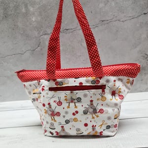 Könnte beinhalten: Eine weiße Stofftasche mit einem wiederkehrenden Muster aus strickenden Hühnern, Wollknäueln, Nadeln und Knöpfen. Die Tasche hat rot-weiß gepunktete Tragegriffe, einen roten Reißverschluss oben und eine horizontale Reißverschlusstasche vorne.