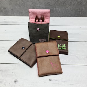 Könnte beinhalten: Eine Sammlung brauner Kunstlederetuis mit rosa und grünen Akzenten. Ein Etui ist geöffnet und zeigt Stricknadeln. Die Etuis haben Knopfverschlüsse und gestickte Details, darunter ein Herz und der Text "Knit HAPPENS".
