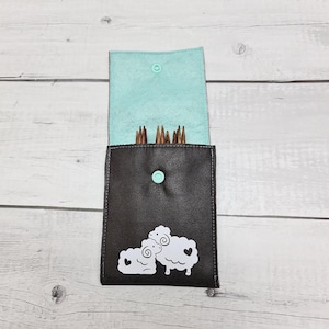Könnte beinhalten: Eine dunkelbraune Stricknadel-Tasche mit hellblauem Innenfutter und einem Druckknopfverschluss. Die Tasche zeigt eine weiße Grafik von zwei Schafen mit herzförmigen Details. Die Tasche ist geöffnet und zeigt mehrere Stricknadeln aus Holz.