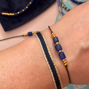 Könnte beinhalten: Zwei Armbänder an einem Handgelenk. Ein Armband ist ein dünnes schwarzes Band mit kleinen goldenen und blauen Perlen. Das andere Armband ist ein breites blaues Band mit goldenen Streifen.