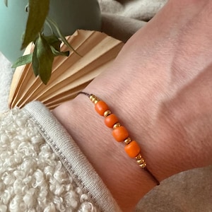 Könnte beinhalten: Ein schlichtes Armband mit braunem Band und orangefarbenen Perlen mit goldenen Akzenten.