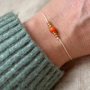 Könnte beinhalten: Ein schlichtes Armband mit einem weißen Band und kleinen goldenen Perlen, akzentuiert mit einer einzigen orangefarbenen Perle.