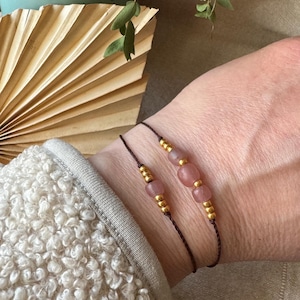 Könnte beinhalten: Zwei zarte Armbänder mit rosa Perlen und goldenen Akzenten auf einem braunen Band. Die Armbänder werden am Handgelenk getragen.