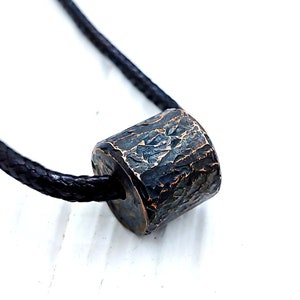 May include: A cylindrical pendant necklace with a textured, dark bronze-colored bead. The bead is suspended on a black cord. The background is white, highlighting the necklace's design and color.