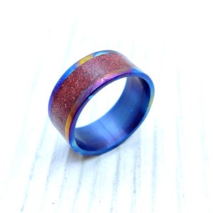 Op de afbeelding: Een brede bandring met een blauw-paarse iriserende afwerking. Het midden van de ring heeft een getextureerde, roodbruine inleg. Een dunne goudkleurige band accentueert de inleg. De ring staat tegen een witte achtergrond.