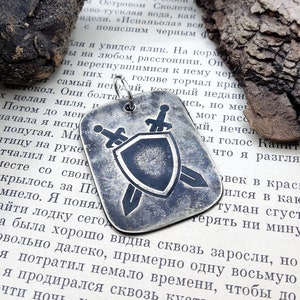 May include: A silver pendant with a shield and two swords design. The pendant is worn and has a distressed look. The pendant is on a book with text in a foreign language.