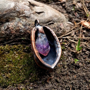 May include: A copper pendant featuring a purple amethyst crystal within. The pendant is shaped like a half-shell and has a rustic, aged appearance.