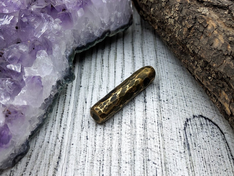 May include: A hammered brass pendant with a textured surface. The pendant is a long, thin cylinder shape with rounded ends.