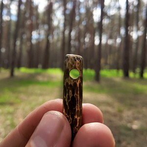 May include: A close-up of a small, hammered copper pendant with a round hole in the center. The pendant is held in a hand against a blurred background of trees.