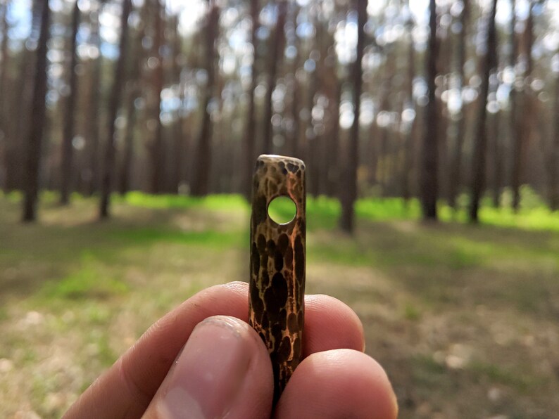 May include: A close-up of a small, hammered copper pendant with a round hole in the center. The pendant is held in a hand against a blurred background of trees.