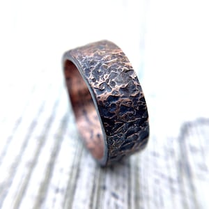 Op de afbeelding: Een brede ring gemaakt van koper, voorzien van een zwaar getextureerd, gehamerd buitenoppervlak met een donkere, geoxideerde patina. De binnenzijde van de ring heeft een gepolijste, metalen koperen afwerking die contrasteert met de donkere textuur.