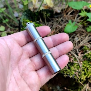 May include: A silver metal lighter with a textured surface and a small loop at the top. The lighter is held in a hand.