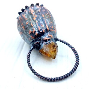 May include: A copper ring with a twisted wire design holds a large, clear, faceted, amber-colored crystal. The crystal is attached to a dark, textured, copper-colored object that resembles a skull.
