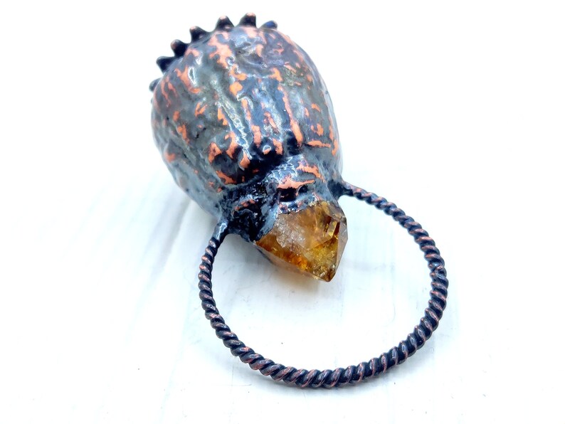 May include: A copper ring with a twisted wire design holds a large, clear, faceted, amber-colored crystal. The crystal is attached to a dark, textured, copper-colored object that resembles a skull.