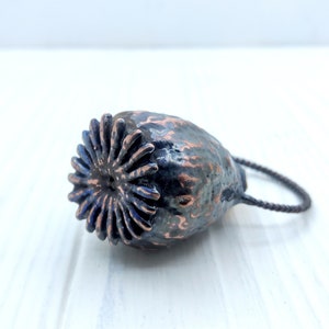 May include: A dark gray and copper colored poppy seed pod pendant with a black cord. The pod has a detailed, textured surface and a circular opening at the top.