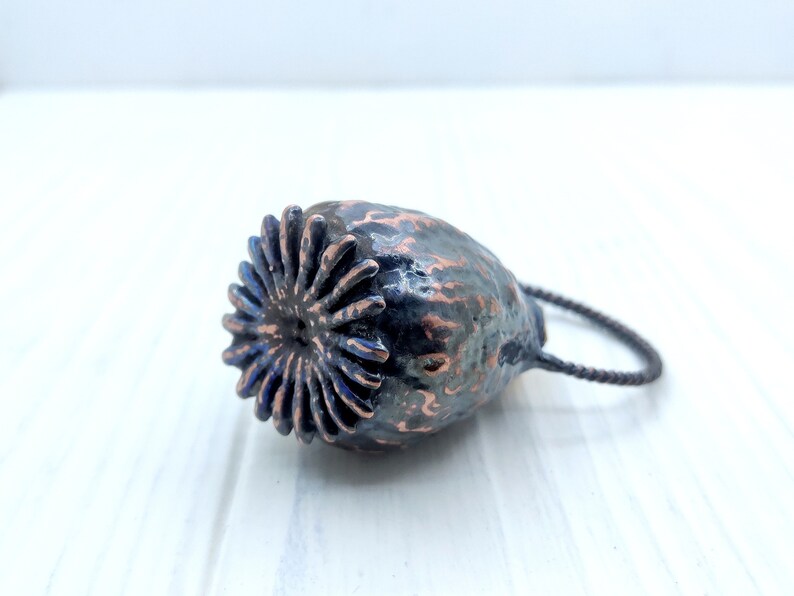 May include: A dark gray and copper colored poppy seed pod pendant with a black cord. The pod has a detailed, textured surface and a circular opening at the top.