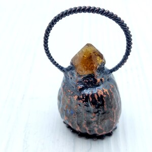 May include: A small, dark brown, textured, ceramic-like object with a gold-colored crystal on top. The object has a twisted wire loop handle.