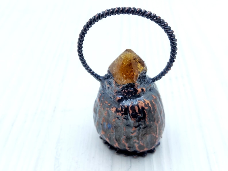 May include: A small, dark brown, textured, ceramic-like object with a gold-colored crystal on top. The object has a twisted wire loop handle.