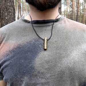 May include: A close-up of a person wearing a gray t-shirt with a black leather cord necklace and a small, natural wood pendant.