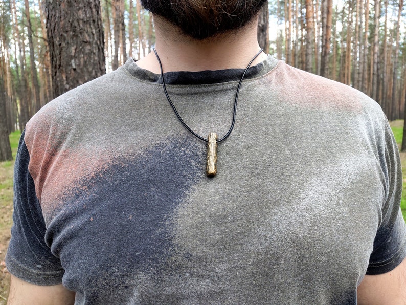 May include: A close-up of a person wearing a gray t-shirt with a black leather cord necklace and a small, natural wood pendant.