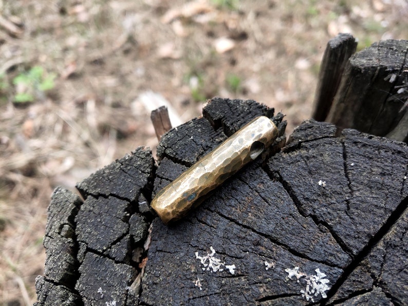 May include: A hammered brass pendant with a hole for a cord or chain. The pendant is a simple, cylindrical shape and has a rustic, handmade look.