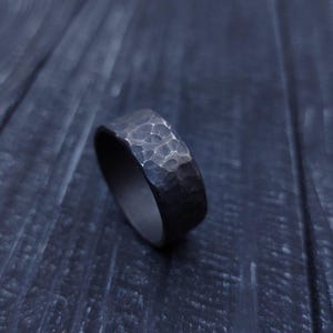 May include: A dark gray ring with a hammered, textured surface. The band features an uneven, dimpled finish across its exterior, creating a rugged appearance. The interior of the ring appears smooth and consistent in color with the outer surface.