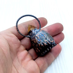 May include: A small, copper-colored pendant with a textured surface and a clear, yellow crystal on top. The pendant is attached to a black, twisted wire loop.