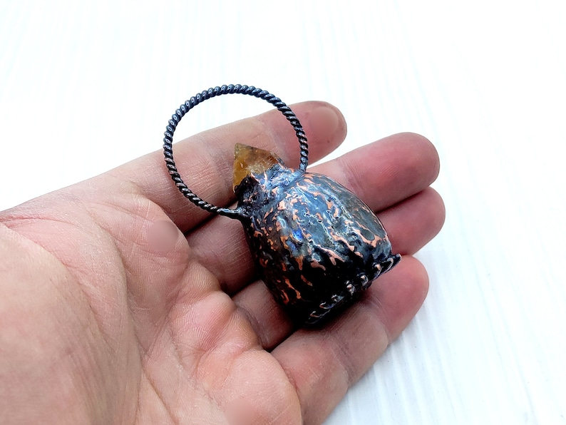 May include: A small, copper-colored pendant with a textured surface and a clear, yellow crystal on top. The pendant is attached to a black, twisted wire loop.