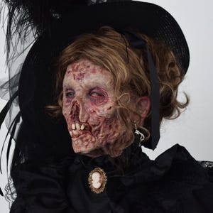 May include: A gruesome zombie head with decaying flesh, wearing a black hat adorned with feathers and a black dress. The zombie has brown hair, a cameo brooch, and a dangling earring. A spooky Halloween decoration.