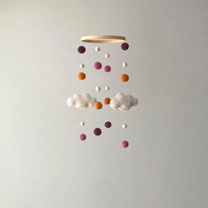 Può includere: Mobile per bambini con un anello di legno e palline di feltro sospese nei toni dell'arancione, viola e bianco. Due soffici nuvole bianche sono sospese sotto le palline, creando una decorazione giocosa.