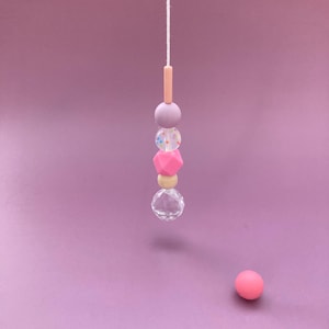 Könnte beinhalten: Ein hängender Perlschmuck mit einer Vielzahl von pastellfarbenen Perlen, darunter rosa, lavendelfarben und klarer Kristall. Eine einzelne rosa Kugel liegt auf der Oberfläche darunter. Die Perlen sind auf einer weißen Schnur vor einem lila Hintergrund aufgefädelt.