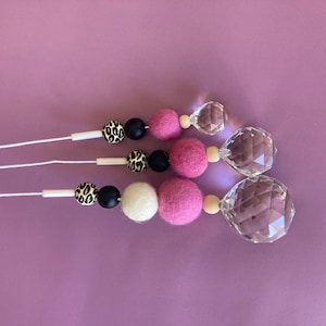 Puede incluir: Tres adornos decorativos con cuentas rosas, blancas, negras y con estampado de leopardo, y colgantes de cristal facetado. Los adornos están ensartados en cordones blancos sobre un fondo morado.