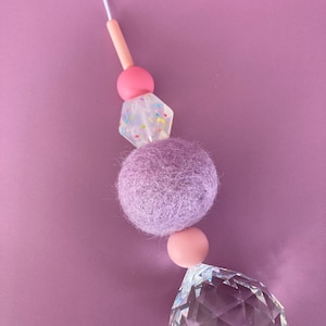 May include: A decorative crystal suncatcher with a string of beads. The beads include a light pink cylinder, a pink sphere, a clear geometric bead with colorful specks, a lavender felt ball, another pink sphere, and a large faceted crystal.