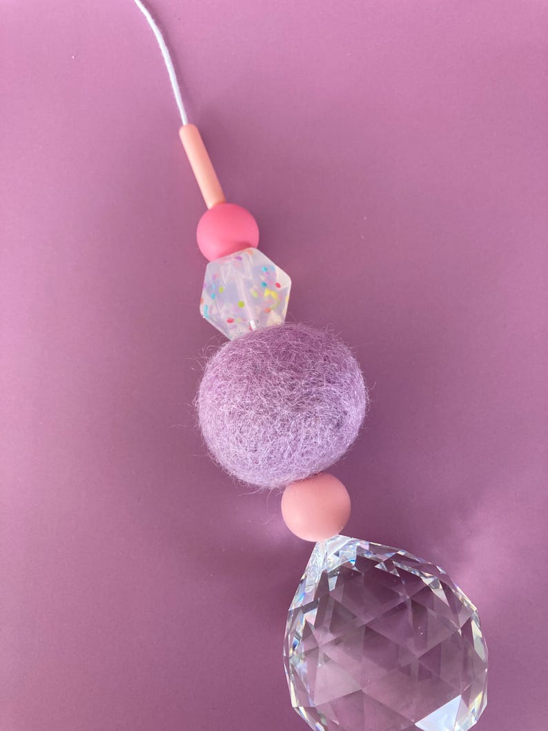 May include: A decorative crystal suncatcher with a string of beads. The beads include a light pink cylinder, a pink sphere, a clear geometric bead with colorful specks, a lavender felt ball, another pink sphere, and a large faceted crystal.