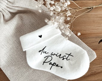 May include: White baby socks with a black heart and the text "du wirst Papa" in black script.