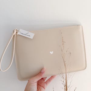 Könnte beinhalten: Beige Clutch mit Handschlaufe und kleinem weißen Herz. Das Etikett trägt die Aufschrift "repsslune". Die rechteckige Tasche ist aus glattem Material gefertigt. Ein schlichtes, elegantes Accessoire.