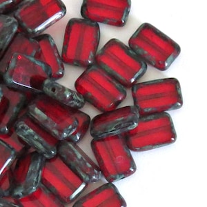 May include: A collection of rectangular glass beads featuring a deep red centre and a dark, mottled green finish along the edges. Each bead has a smooth, polished surface and a central hole, arranged in a scattered pile on a plain white surface.