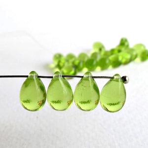 May include: Close-up of four translucent, teardrop-shaped beads strung on a wire. The beads are a vibrant, light green color. A cluster of smaller, round green beads is out of focus in the background. The beads are likely for jewelry making.