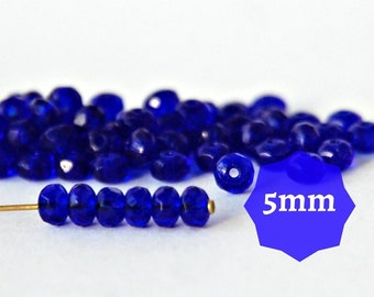 May include: A close-up of a pile of small, cobalt blue glass beads. The beads are faceted and have a shiny finish. A single strand of beads is laid out on a white surface with a gold wire running through the beads. The text "5mm" is displayed in a blue circle.