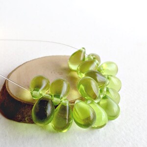 May include: A collection of translucent, olive-green glass beads in a teardrop shape, strung on a thin, clear wire. The beads are arranged on a small, round, wooden slice, with a white background.