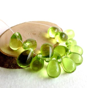 May include: A collection of olive green glass beads in various shapes, including teardrops and spheres, strung on a clear cord. The beads are displayed on a natural wood slice, creating a contrast of textures and colors. The beads have a translucent quality.