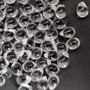 May include: A group of clear teardrop-shaped crystal beads with a carved design. The beads are scattered on a black surface.