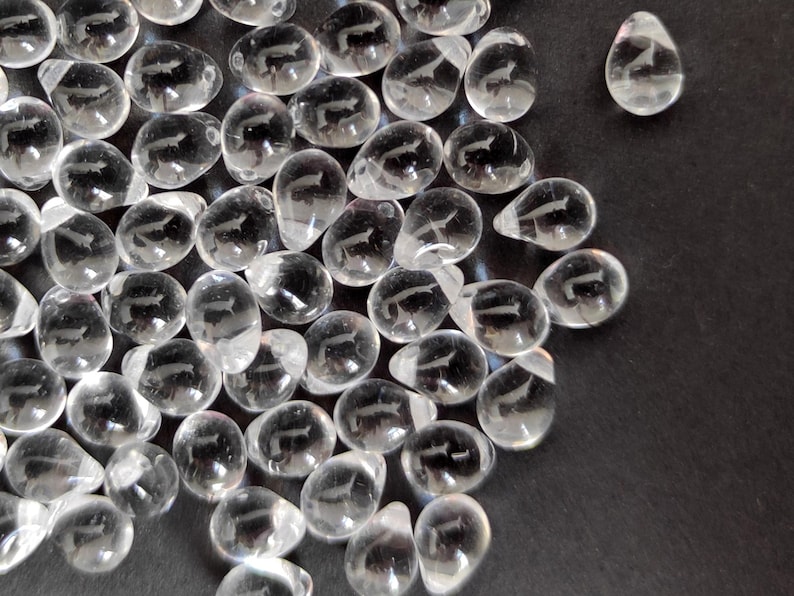 May include: A group of clear teardrop-shaped crystal beads with a carved design. The beads are scattered on a black surface.
