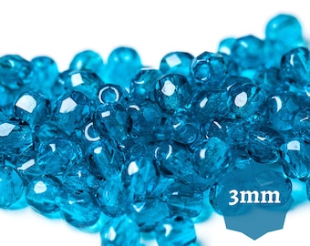 May include: A close-up of a pile of small, turquoise blue glass beads. The beads are 3mm in size and have a faceted, crystal-like appearance.