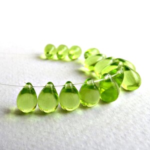 May include: A strand of translucent, teardrop-shaped beads in a vibrant, light green hue. The beads are strung on a thin, clear wire, with several in focus and others blurred in the background. The beads are evenly spaced and appear to be made of glass.