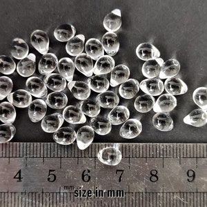 May include: A pile of clear glass teardrop beads, measuring approximately 6 mm in size, against a black background. A ruler is included for scale.