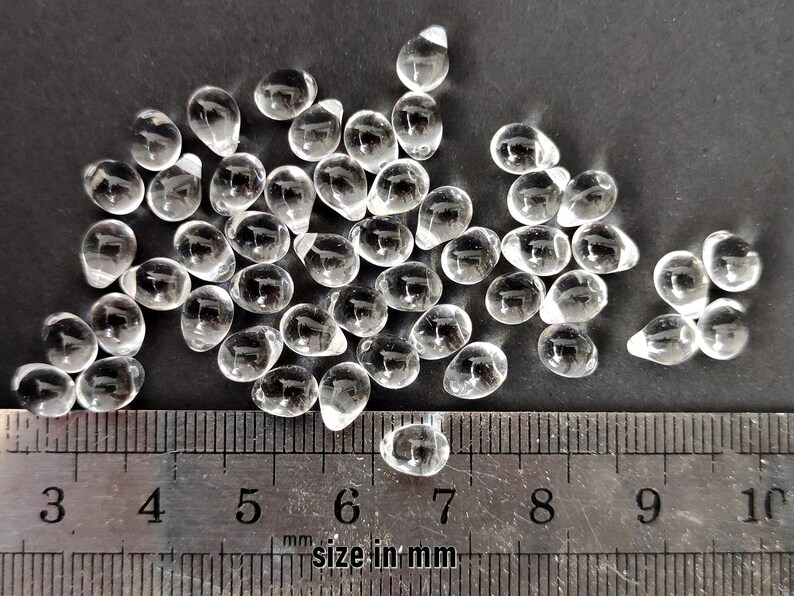 May include: A pile of clear glass teardrop beads, measuring approximately 6 mm in size, against a black background. A ruler is included for scale.