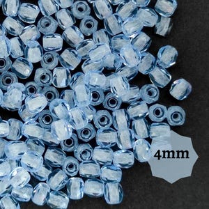 Pode incluir: Um close-up de um monte de contas de vidro azul claro. As contas medem 4 mm e têm uma forma quadrada facetada.