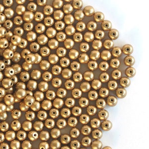 以下が含まれることがあります： 小さな丸い金色のビーズのコレクション。各ビーズには中央に小さな穴があります。ビーズは白い表面に散らばっており、テクスチャード加工された外観を作り出しています。ジュエリー作りや工芸に最適です。